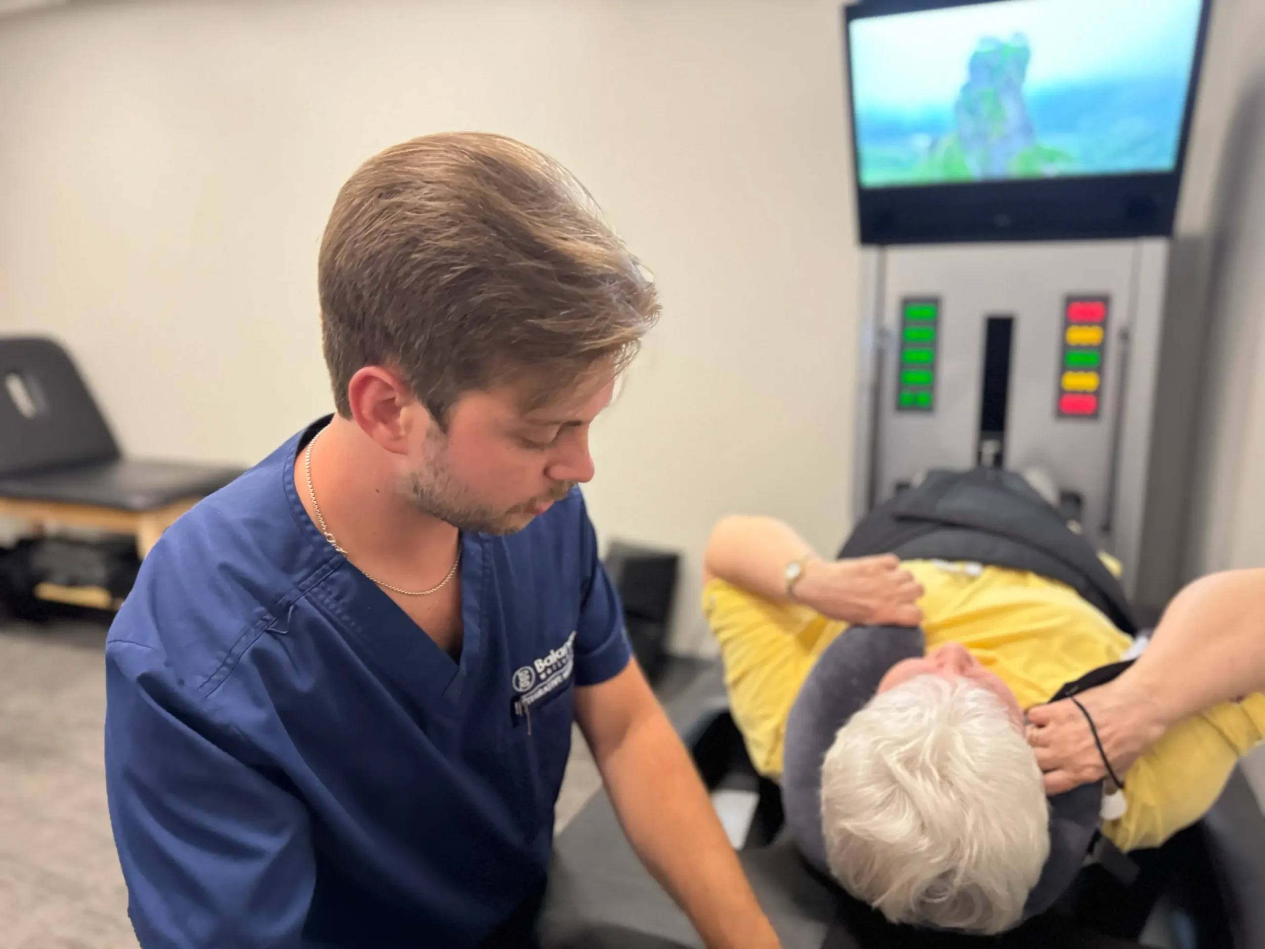 Healthcare practitioner adjusting an older patient lying on a therapy or rehabilitation machine with a screen in the background.